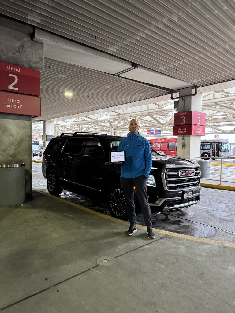Cameron in the VME jacket with a name sign beside the SUV at Denver Airport, Level 5, Island 2, Limo Section E